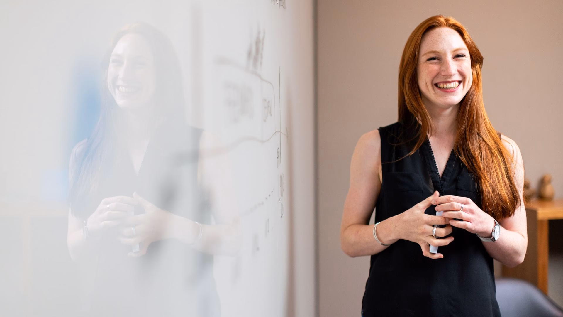 "A smiling woman with long red hair stands in front of a whiteboard, delivering a presentation. Her reflection is visible on the glass surface, adding depth to the image. She appears confident and engaged, conveying enthusiasm in a professional setting.